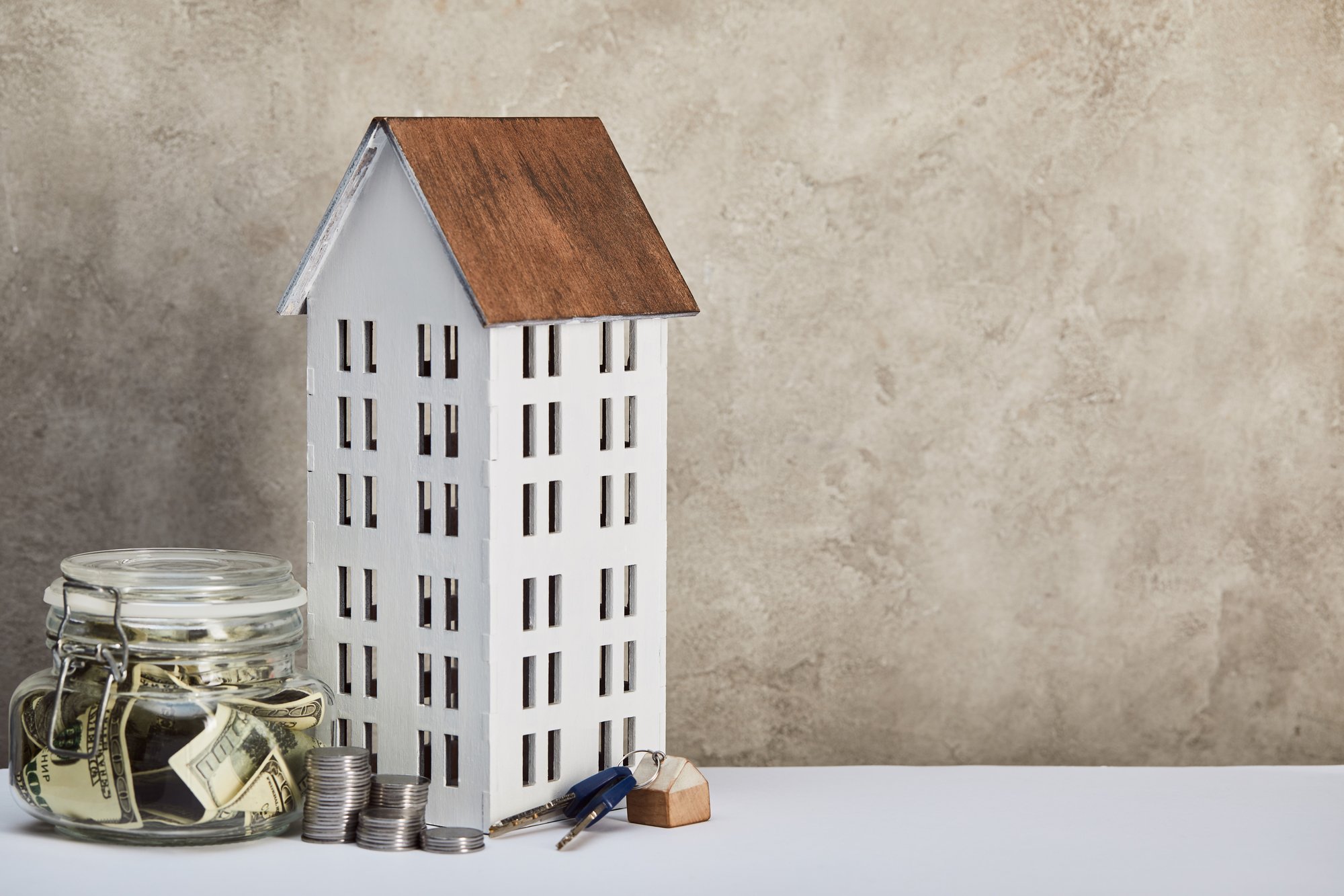 House model, moneybox with dollar banknotes, keys and silver coins on white table on grey background, real estate concept